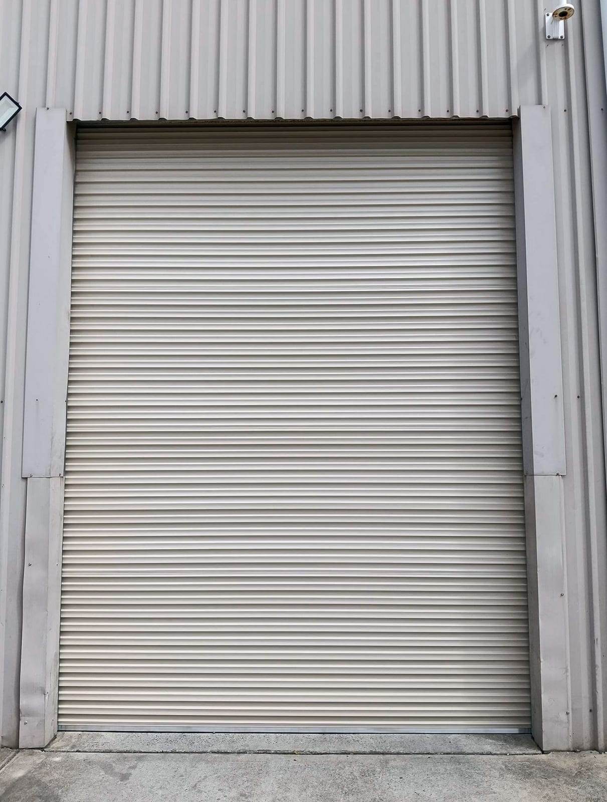 Tall beige rolling steel door, closed, set into the ribbed metal wall of a commercial building above a concrete apron
