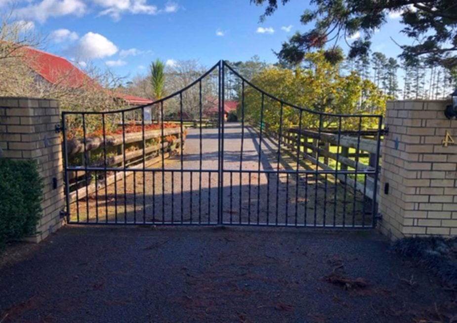 Ornamental wrought iron double swing driveway gate with an arched top, hung between brick pillars at the end of a gravel drive