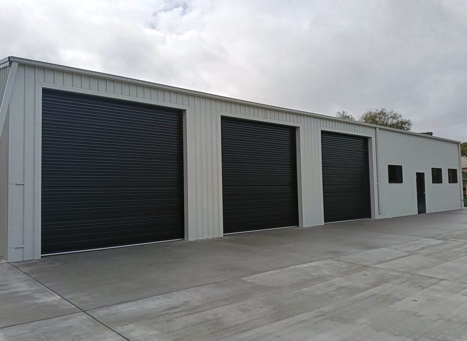 Three charcoal roll-up doors side by side in a new light gray steel workshop building with a wide concrete apron