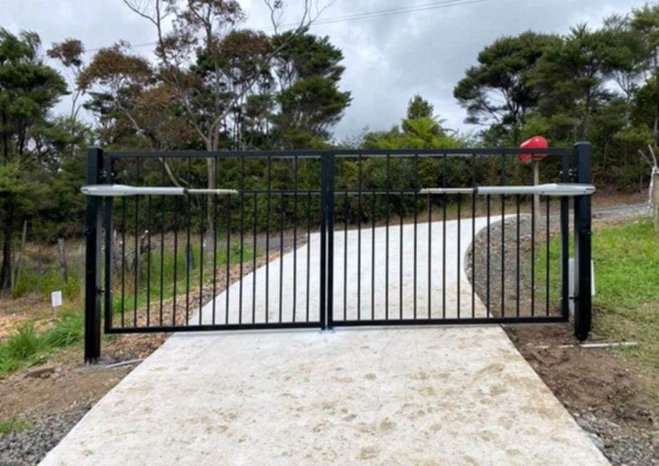 Black steel picket double swing gate fitted with two swing-arm gate openers, on a new concrete apron leading to a gravel driveway