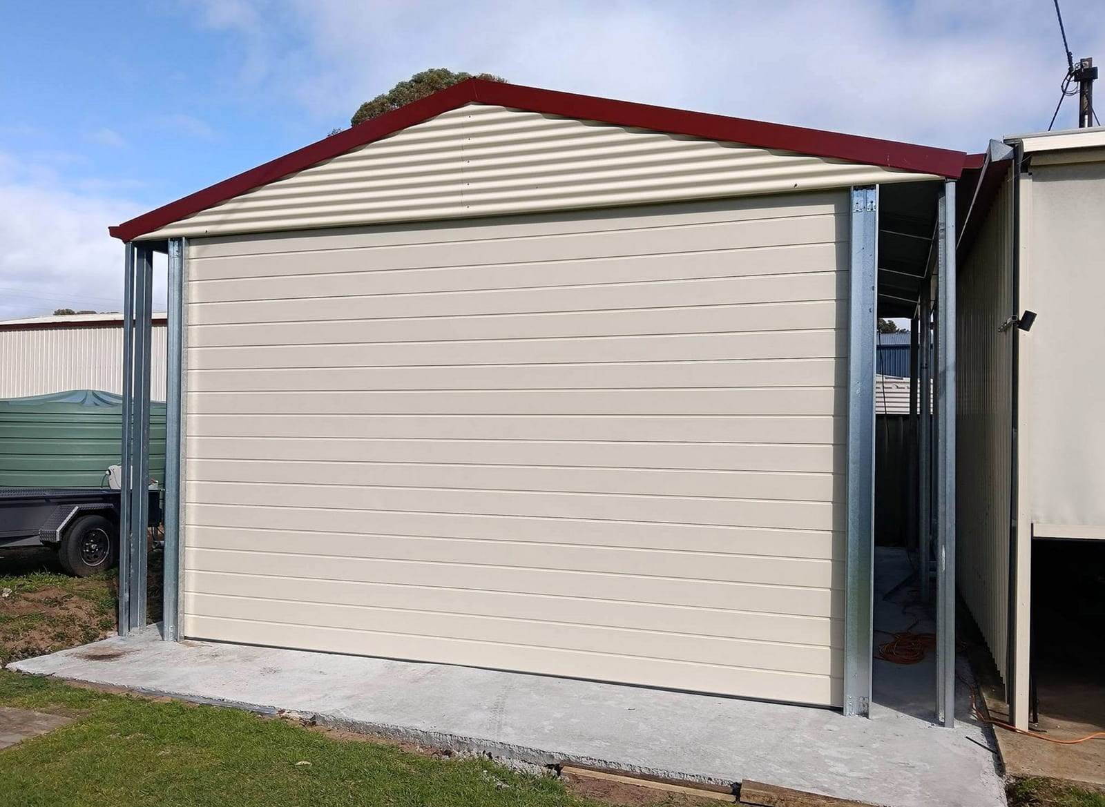 Cream ribbed steel garage door, closed, in a new steel-framed garage with dark red roof trim, standing on a fresh concrete slab