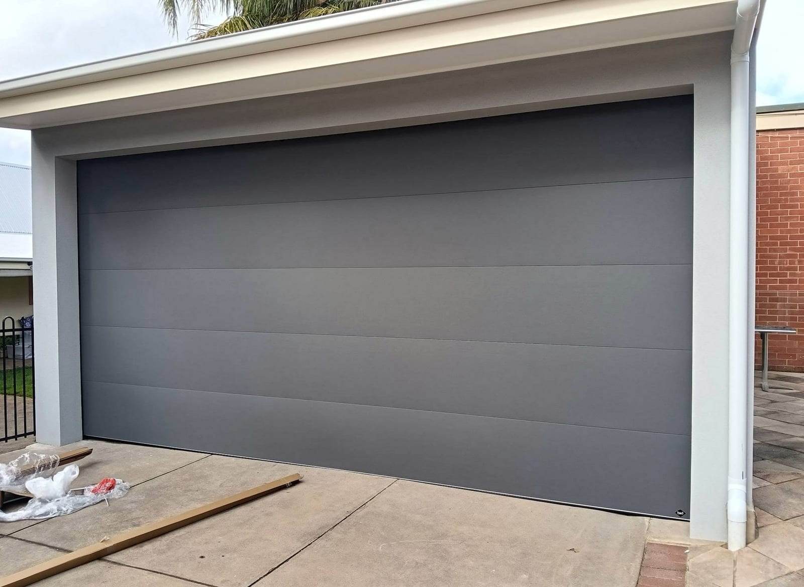 Wide gray flush-panel sectional garage door in a stucco opening, with packing plastic and a wooden board still on the driveway after the install