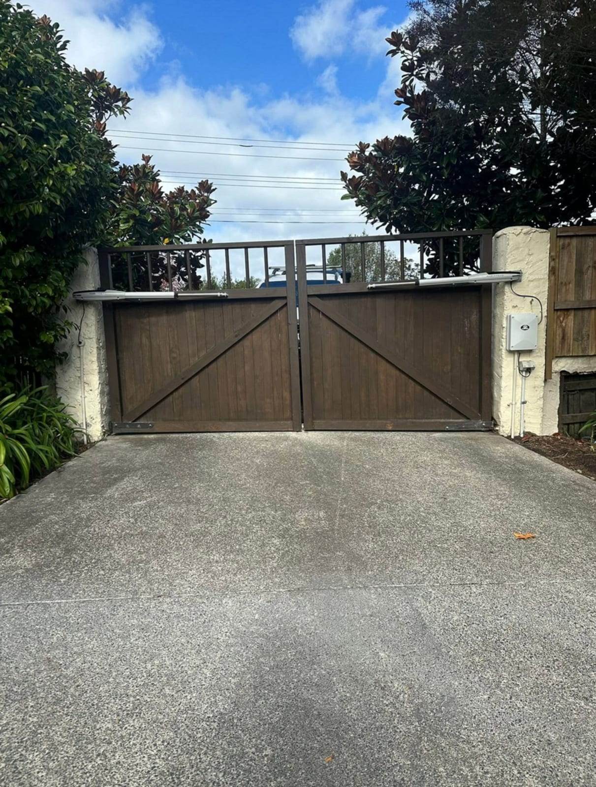 Dark-stained wood double swing driveway gate with a metal picket top rail, automated with two articulated arm gate openers mounted on stucco pillars