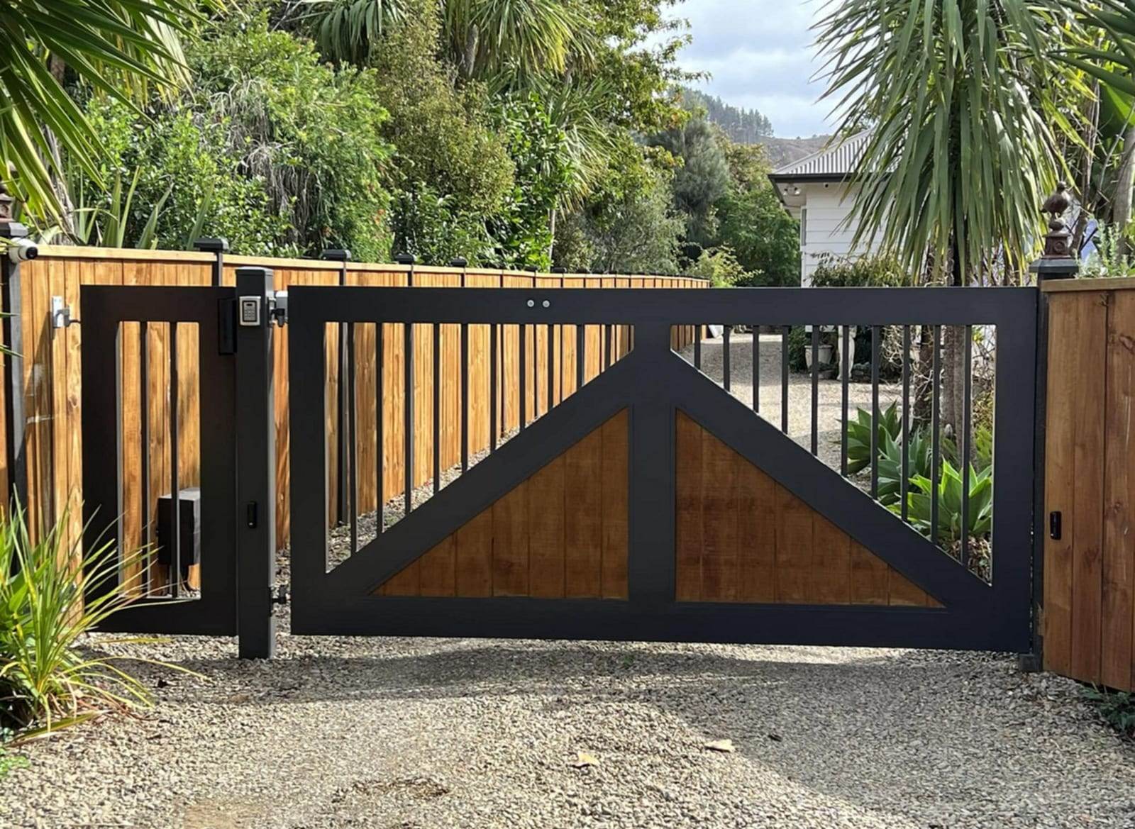Black steel-framed double swing gate with triangular wood infill panels and vertical bars, with a keypad post beside a gravel driveway