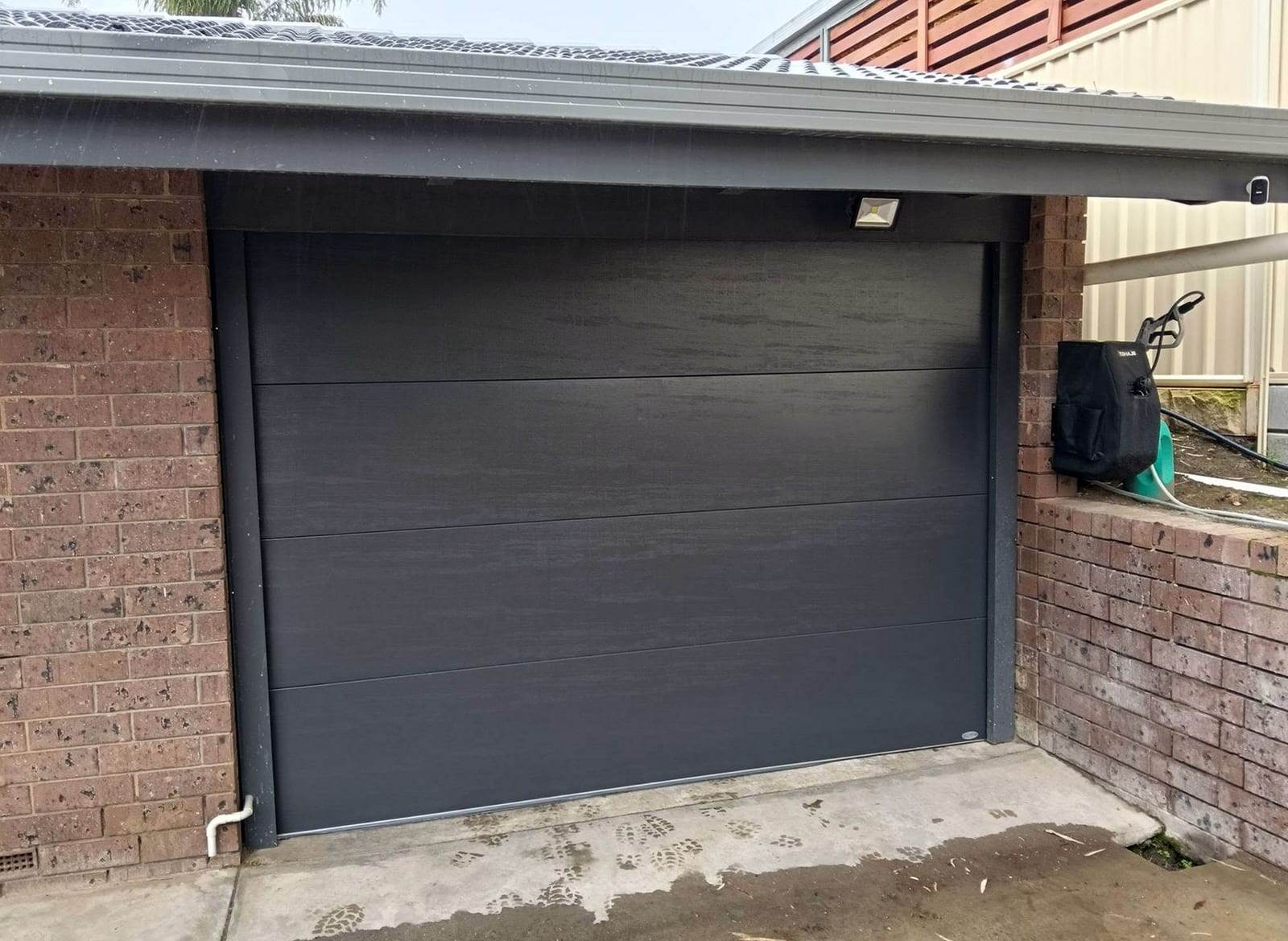 Charcoal sectional garage door with a woodgrain texture and four flush panels, in a brick garage with a floodlight above the opening
