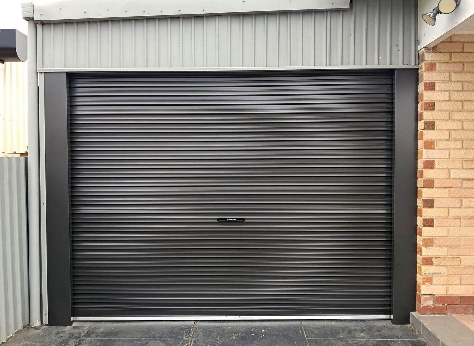 Matte black roll-up garage door between black jamb covers, in a steel-clad wall next to a brick house