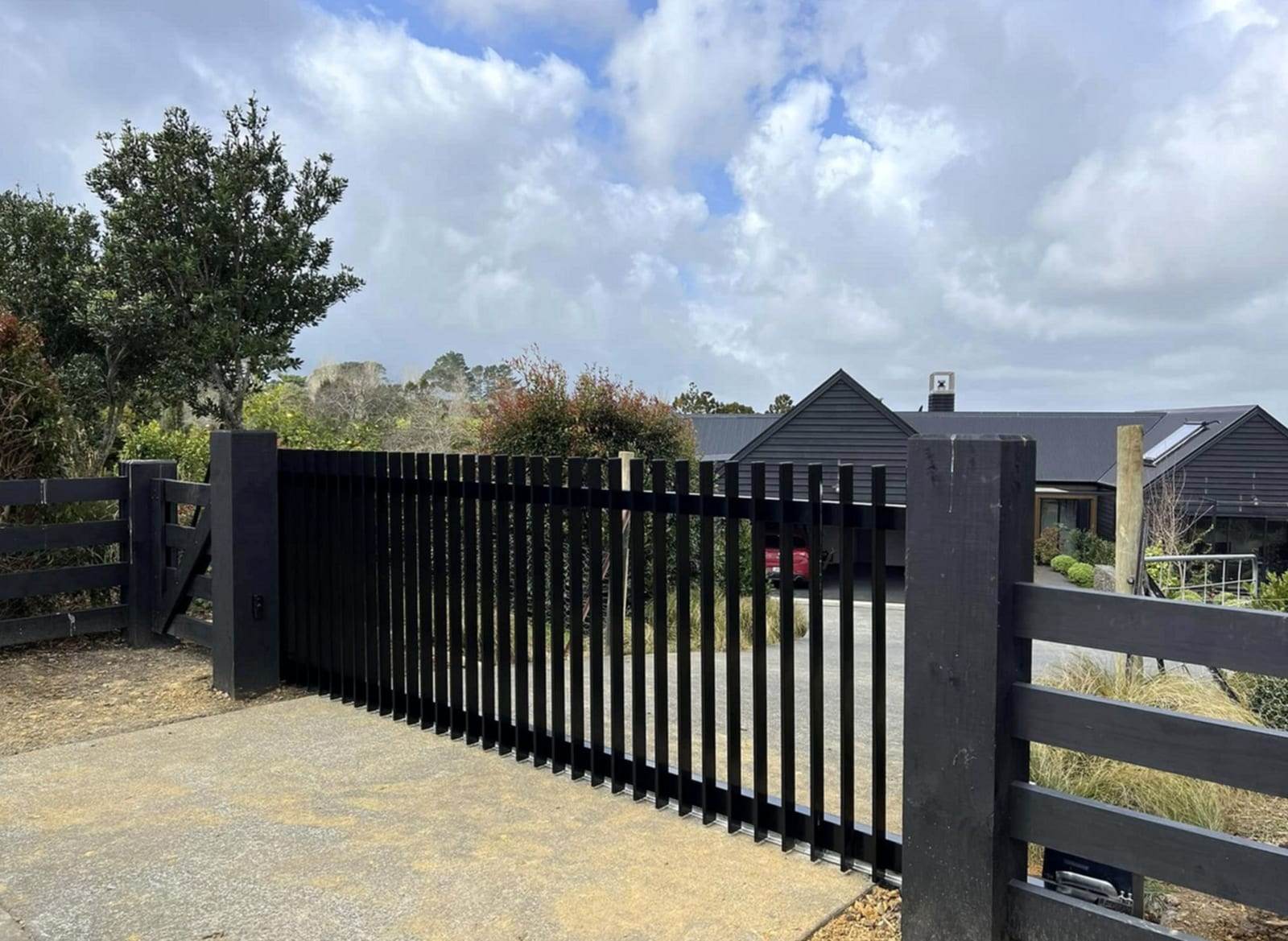 Black vertical-slat aluminum sliding driveway gate between black timber posts, with a modern house and carport behind it
