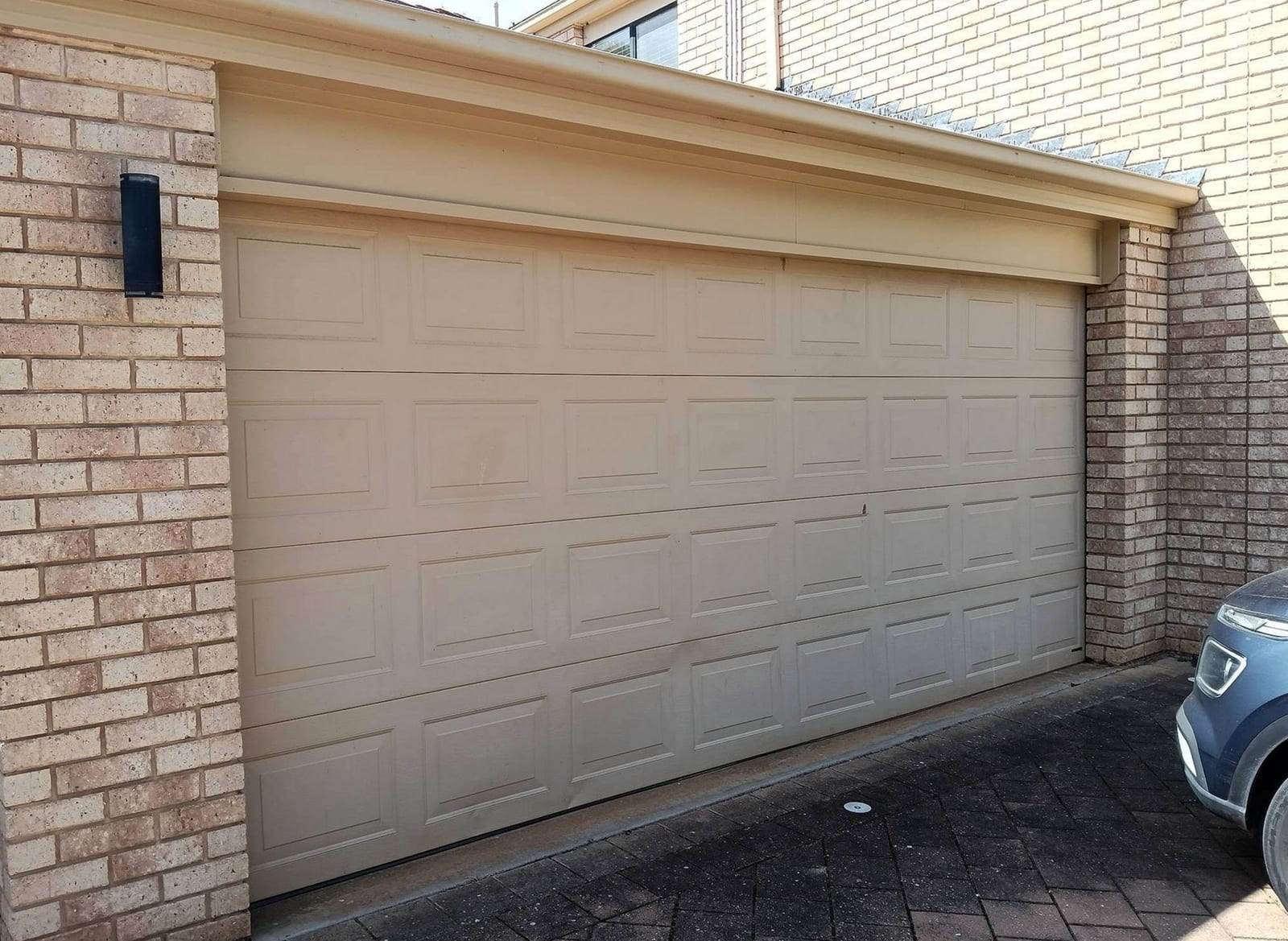 Beige raised-panel double garage door with rows of short panels, set in a tan brick two-car garage, with the front of a parked car at the right
