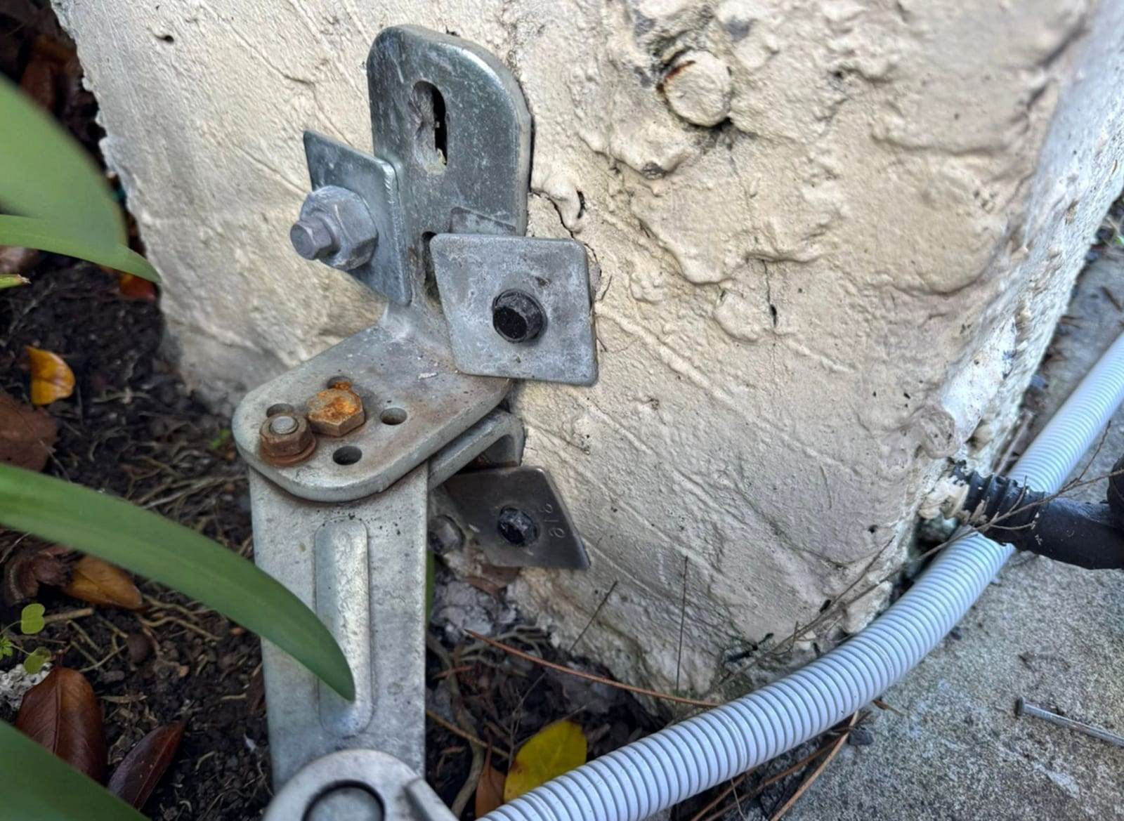 Close-up of a galvanized adjustable gate hinge and mounting bracket bolted to a stucco pillar, with flexible conduit for the operator wiring