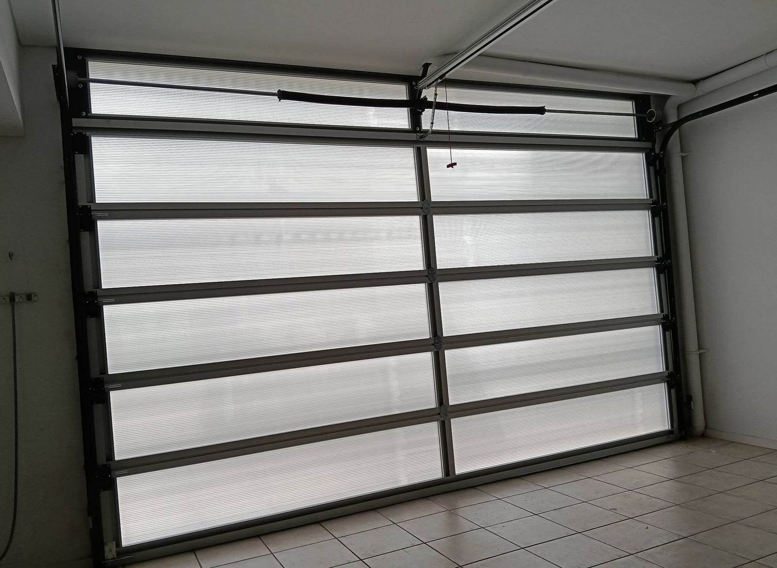 Inside view of a full-view aluminum garage door: two torsion springs on the shaft above the door, the opener rail running to the ceiling, the red emergency release cord and the curved track at the side